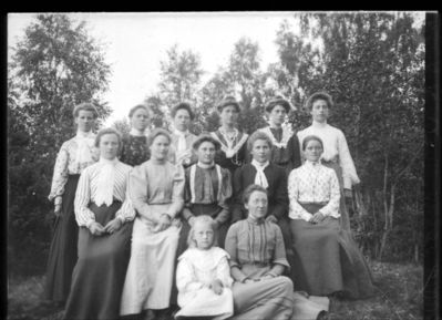 Gruppebilde  1900 - 10
Elleve unge kvinner sittende oppstilt. Kvinne og liten pike sittende foran. Utendørs sommer.
Nøkkelord: gruppe;kvinner;dame;barn;1900
